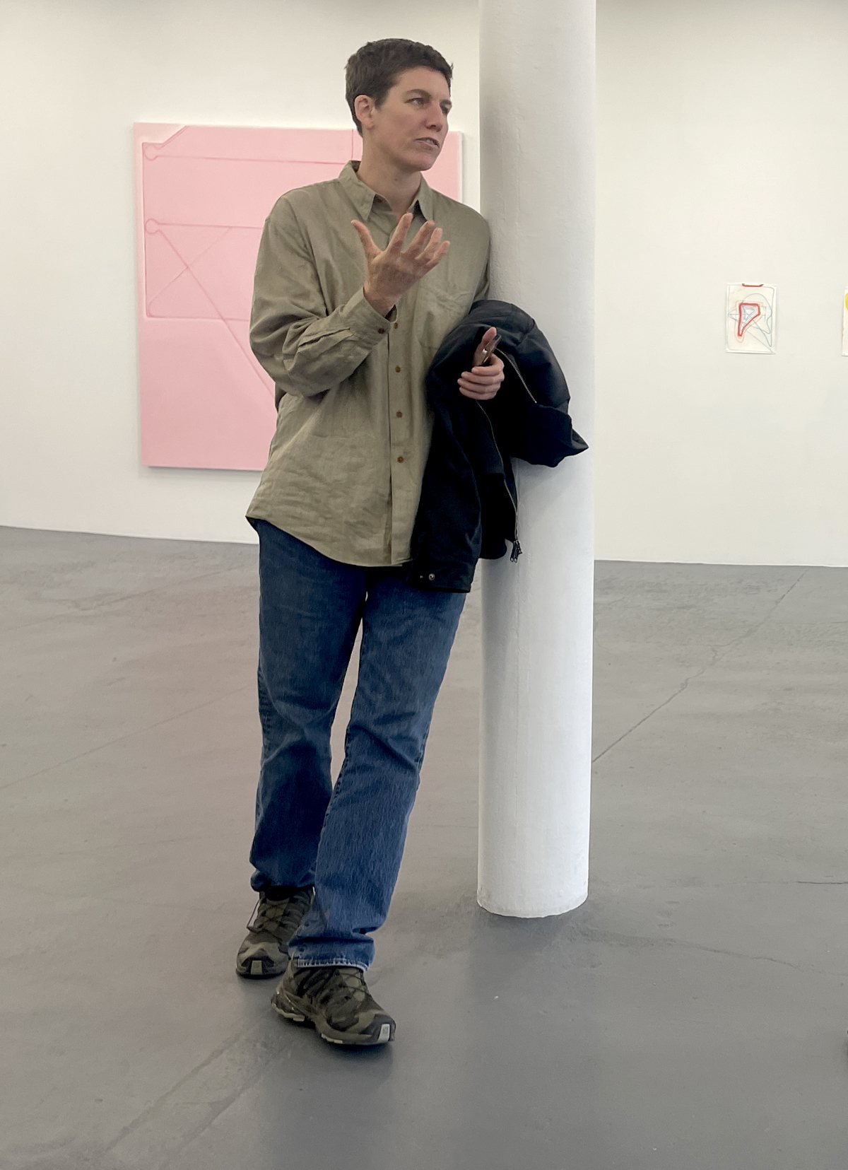 Fox Hysen speaking, standing beside a column in a gallery with paintings on the wall behind her.